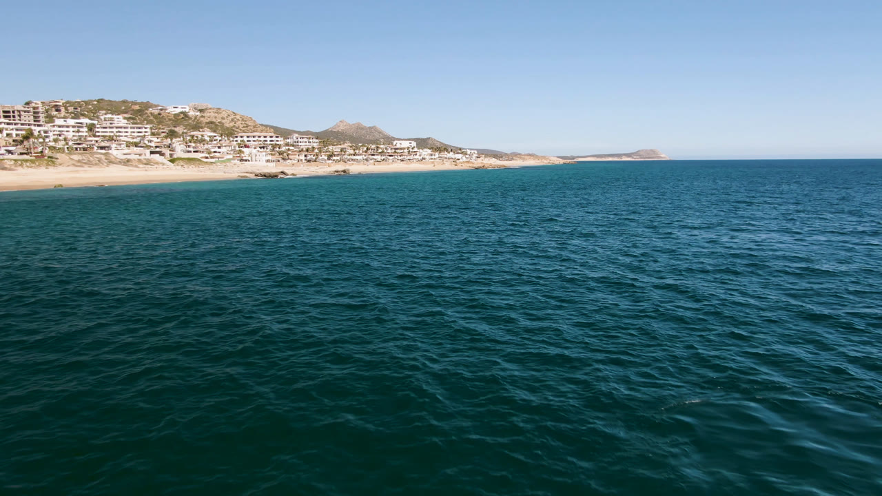 Opening frame of Laguna Beach Los Cabos with blue-green ocean in the foreground, sandy coast and hillside buildings at left, distant rocky landforms, and no visible people.
