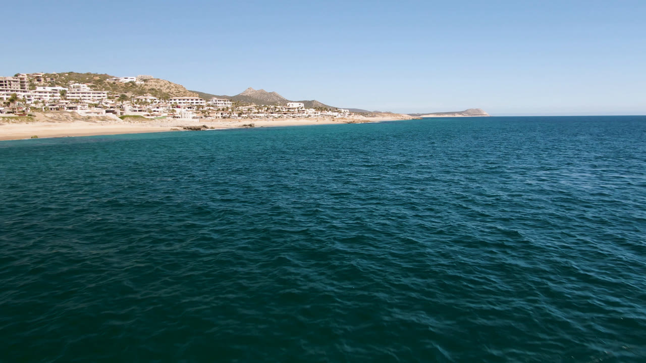 Second frame over Laguna Beach Los Cabos showing textured ocean water across the foreground, bright sand and white hillside buildings to the left, distant rocky coast, and no visible people.