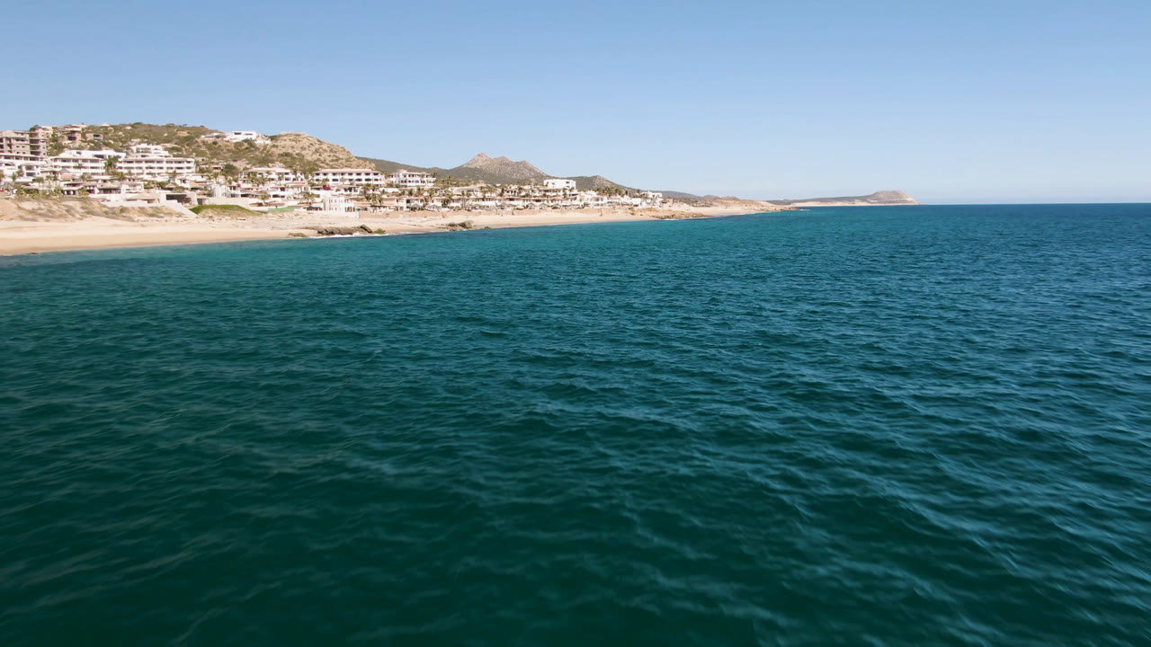 Opening frame of Laguna Beach Los Cabos with blue-green ocean in the foreground, a sandy beach at left, hillside buildings beyond it, and no clearly visible people.