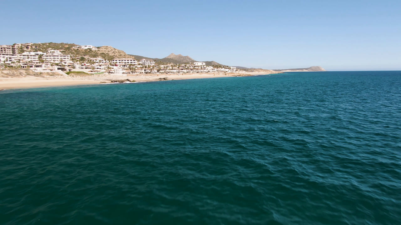 Second frame from Laguna Beach Los Cabos showing rippled ocean water, the beach and white hillside buildings slightly left of center, and no clearly visible people.