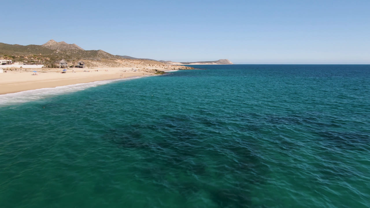 Opening frame of Laguna Beach Los Cabos with turquoise water in the foreground, pale sand at left, low hills beyond the shore, and no visible people.