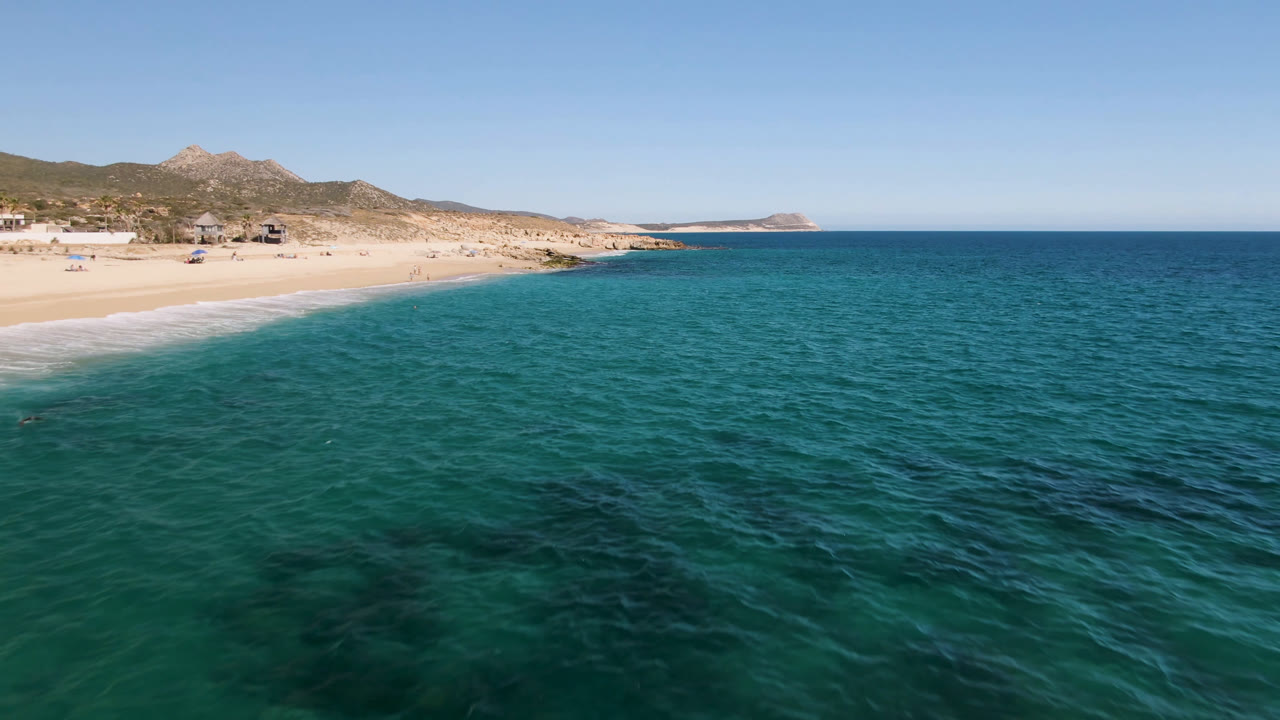 Laguna Beach Los Cabos frame with rippled blue-green water dominating the foreground, surf touching the sand at left, low hills behind the beach, and no visible people.