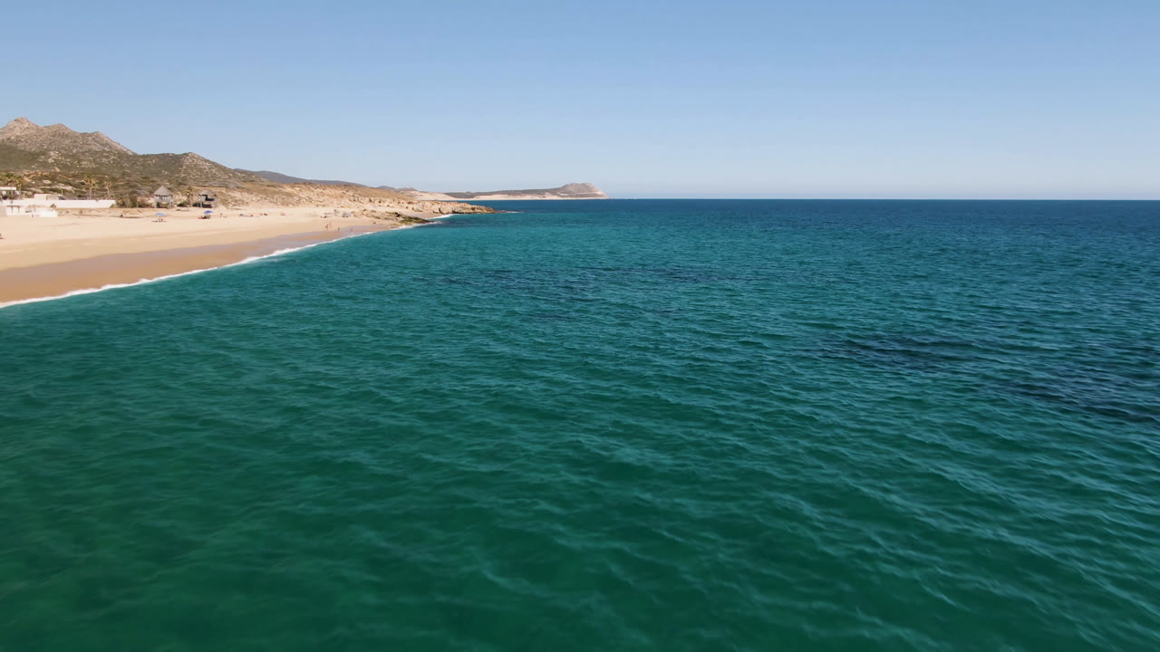 Wide aerial view of Laguna Beach in Los Cabos, Mexico, showing textured turquoise water, a pale sand shoreline, low hills, and no visible people.