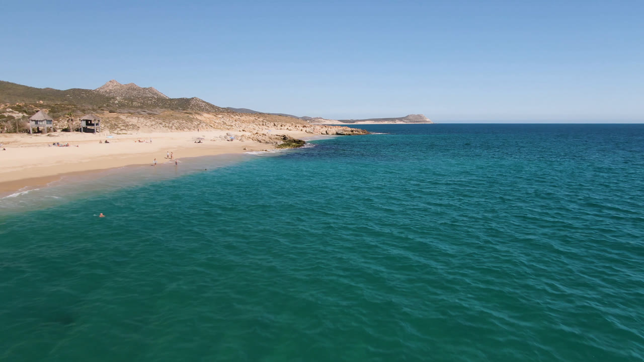 Aerial view of turquoise ocean beside a sandy beach and dry hills.
