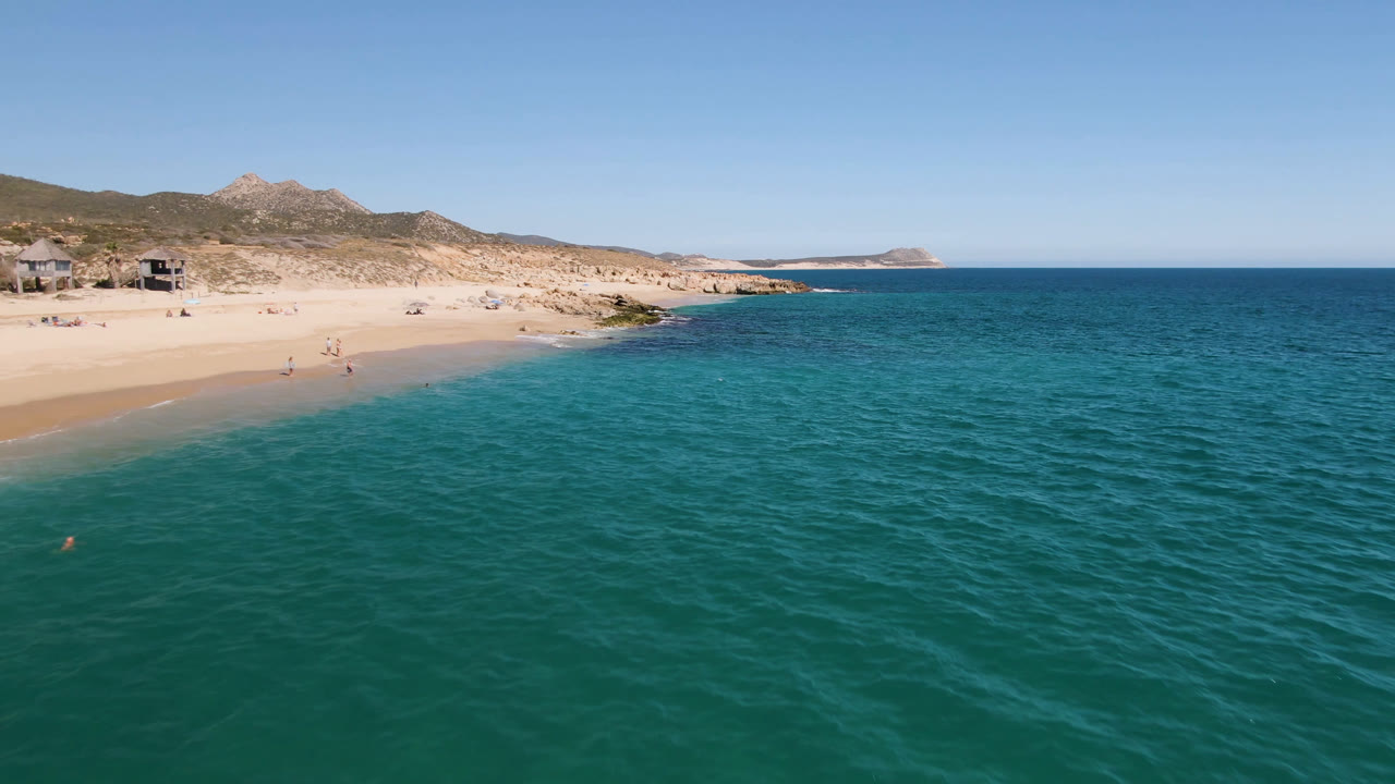 Laguna Beach Los Cabos appears from above with the beach running along the left edge, rippled turquoise water across the foreground, small people on the sand, and low mountains behind.
