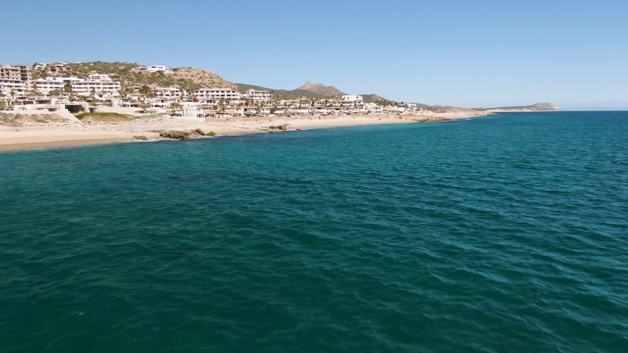 A broad aerial frame over Laguna Beach Los Cabos water shows small ripples in the foreground, shoreline at upper left, and hillside buildings beyond, with no visible people.