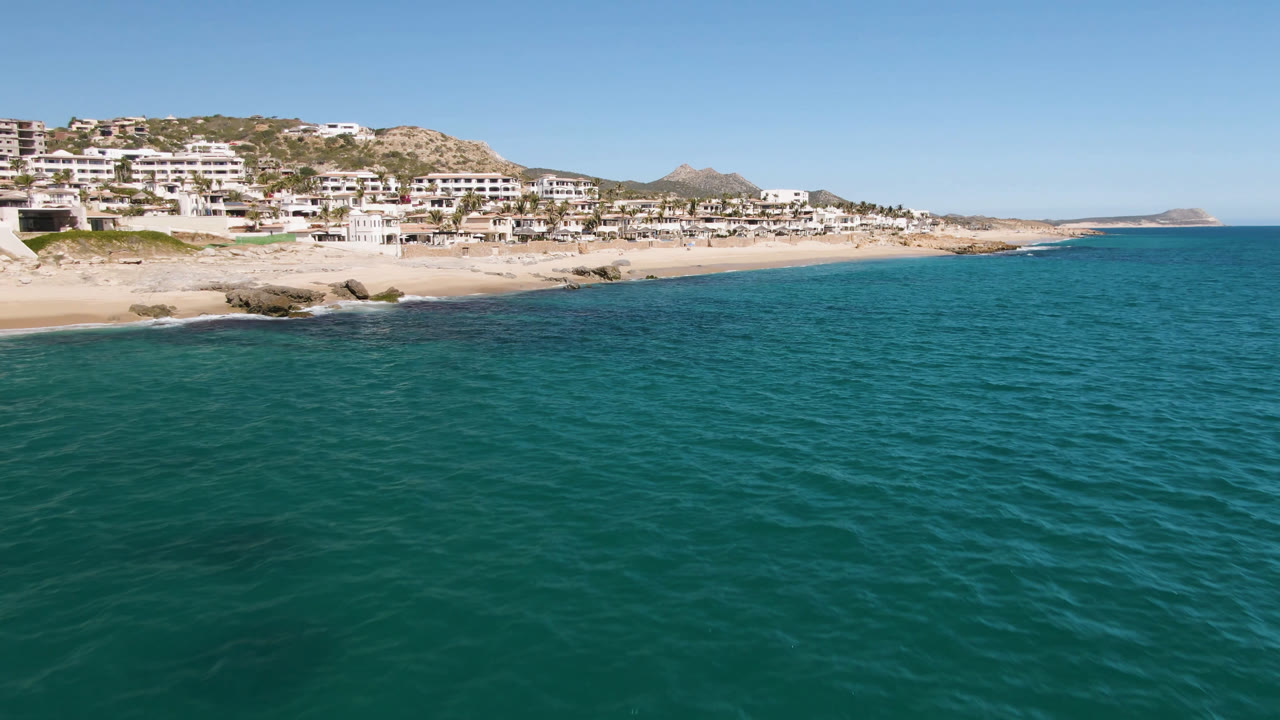 Opening frame over Laguna Beach Los Cabos shows turquoise water in the foreground, a sandy and rocky shoreline, hillside houses, clear sky, and no visible people.