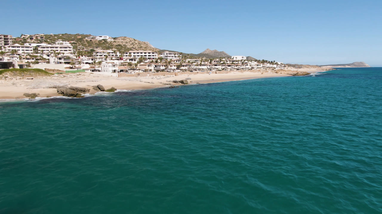 At 0:01, Laguna Beach Los Cabos remains framed from above with teal water, pale sand, shoreline rocks, white hillside homes, and no visible people.