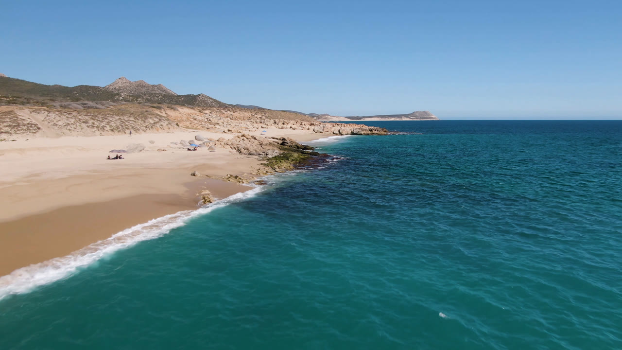Aerial view of Laguna Beach Los Cabos with turquoise water in the foreground, pale sand to the left, rocky shoreline near center, distant hills, and small beachgoers visible on the sand.