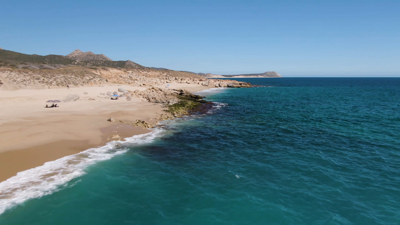 A wide aerial frame at Laguna Beach Los Cabos shows a wave line curling onto the beach, dark rocks along the shore, blue water to the right, and small distant people near umbrellas.