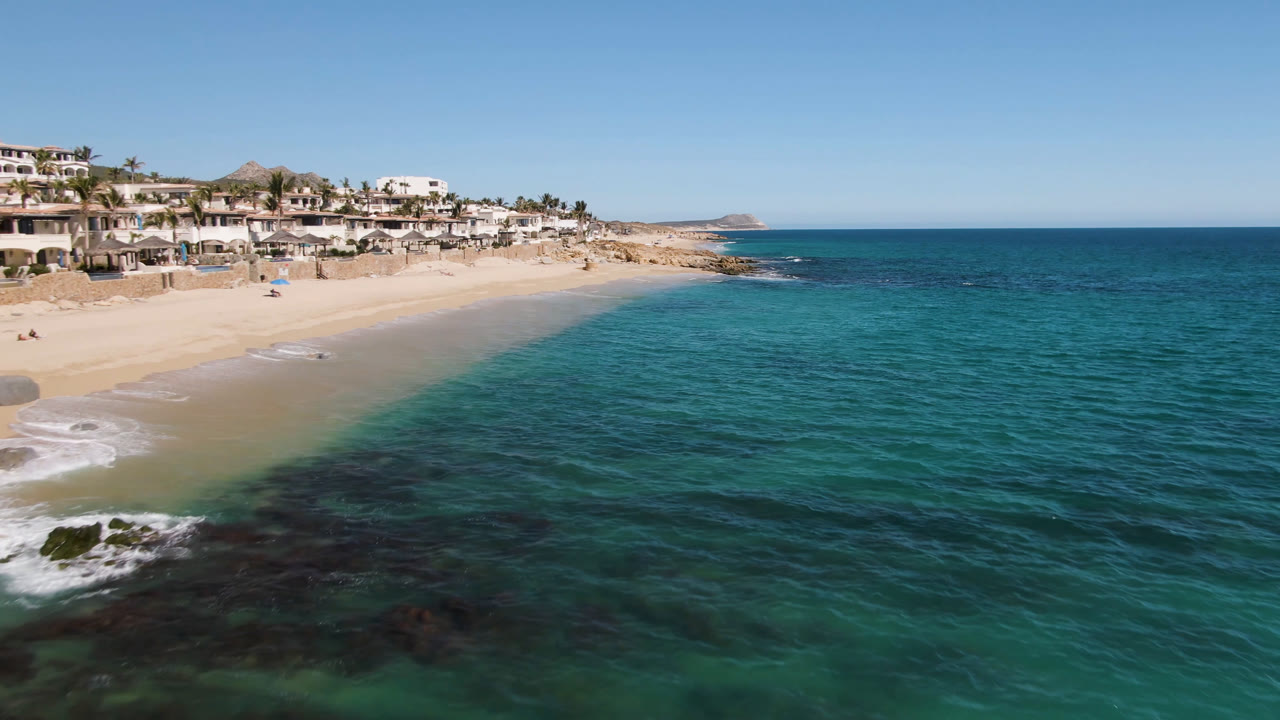 Aerial view of turquoise ocean beside a sandy beach and hillside houses.