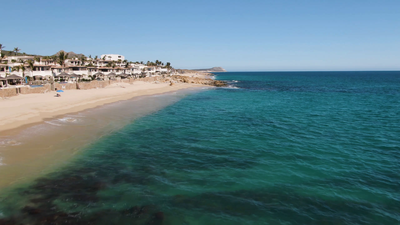 Aerial frame of Laguna Beach Los Cabos with more open water across the foreground, a narrow sand edge at left, white houses above the beach, and no visible people.