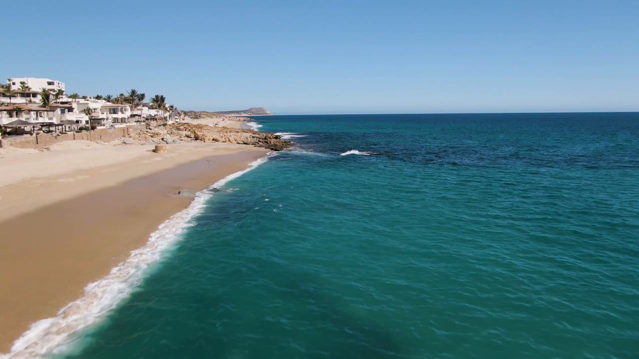 Aerial frame of Laguna Beach Los Cabos showing a curved sandy beach at left, turquoise ocean at right, rocky coast, white buildings, and no visible people.