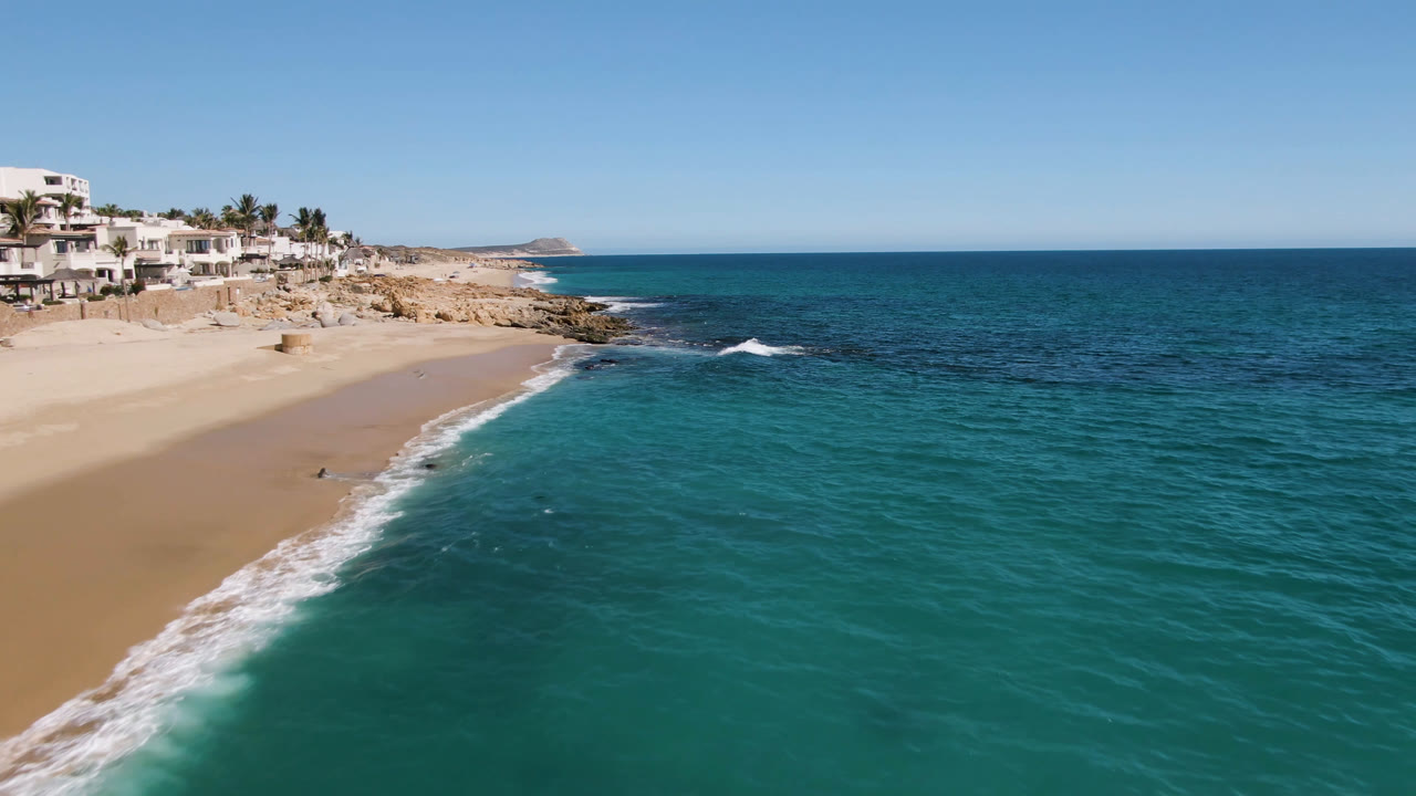Aerial view of Laguna Beach Los Cabos with a white wave line along the sand, turquoise water in the foreground, rocky shore, white buildings, and no visible people.