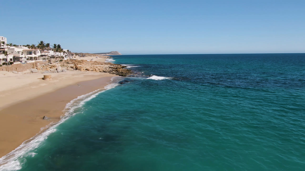 Drone view over clear blue water beside sand, rocks, and coastal buildings