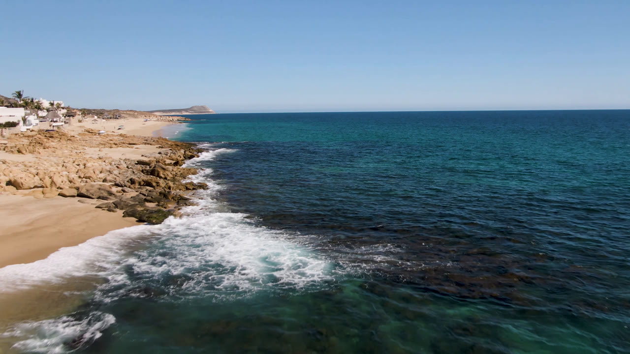 Laguna Beach Los Cabos shoreline seen from above, with waves foaming over rocks beside the sand, open blue water to the right, distant coast ahead, and no visible people.