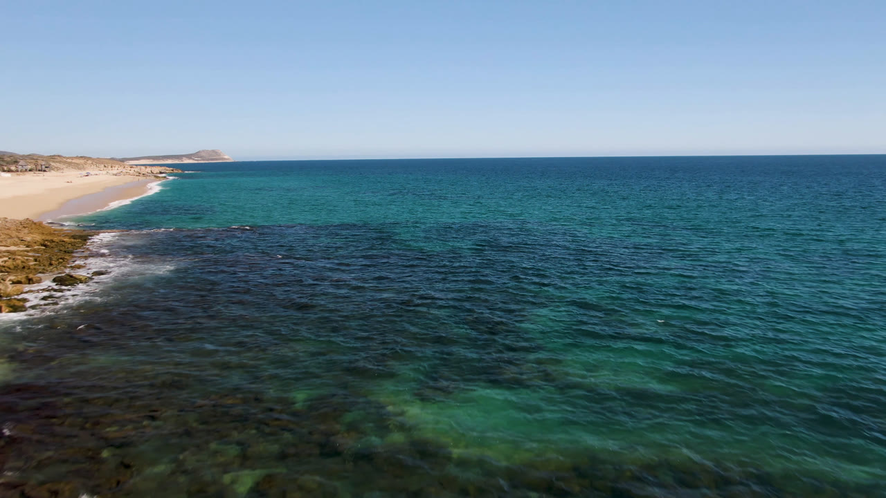 Aerial view at Laguna Beach in Los Cabos with dark nearshore reef in the foreground, turquoise ocean beyond, sand beach at left, distant low hills, and no visible people.