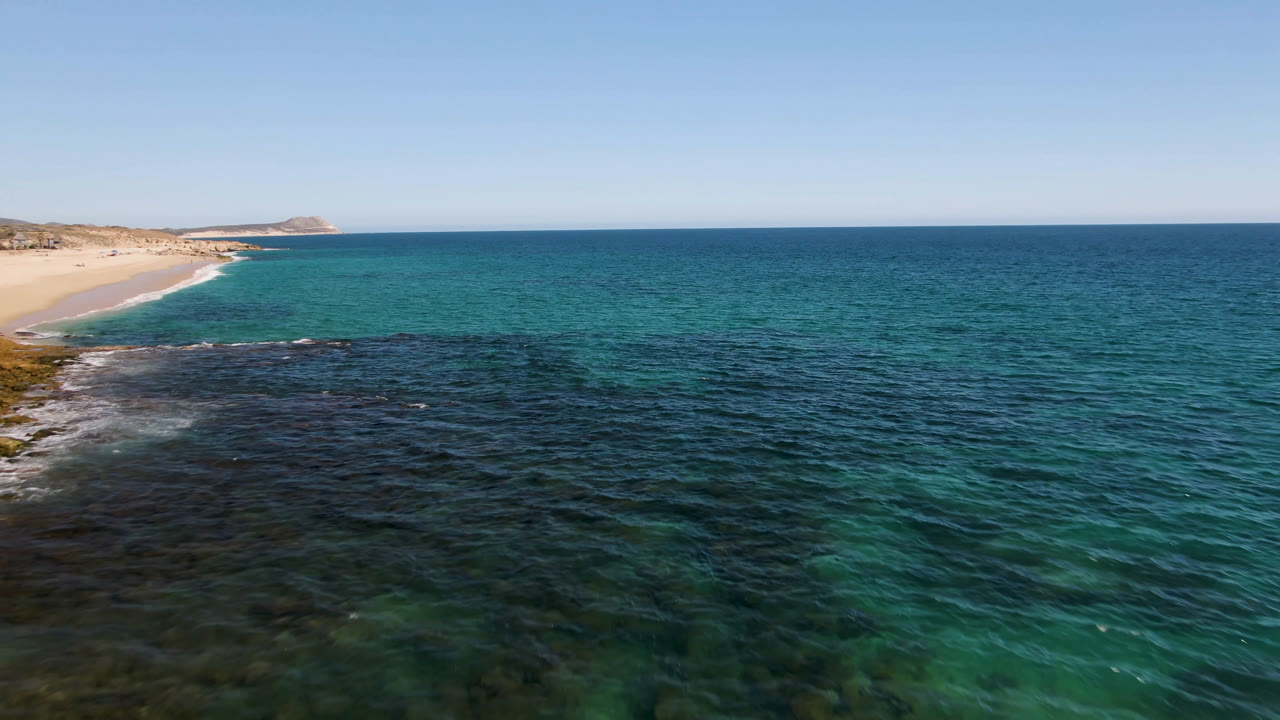 Laguna Beach in Los Cabos appears from above with blue-green water covering most of the frame, dark reef across the lower foreground, pale beach on the left, and no visible people.