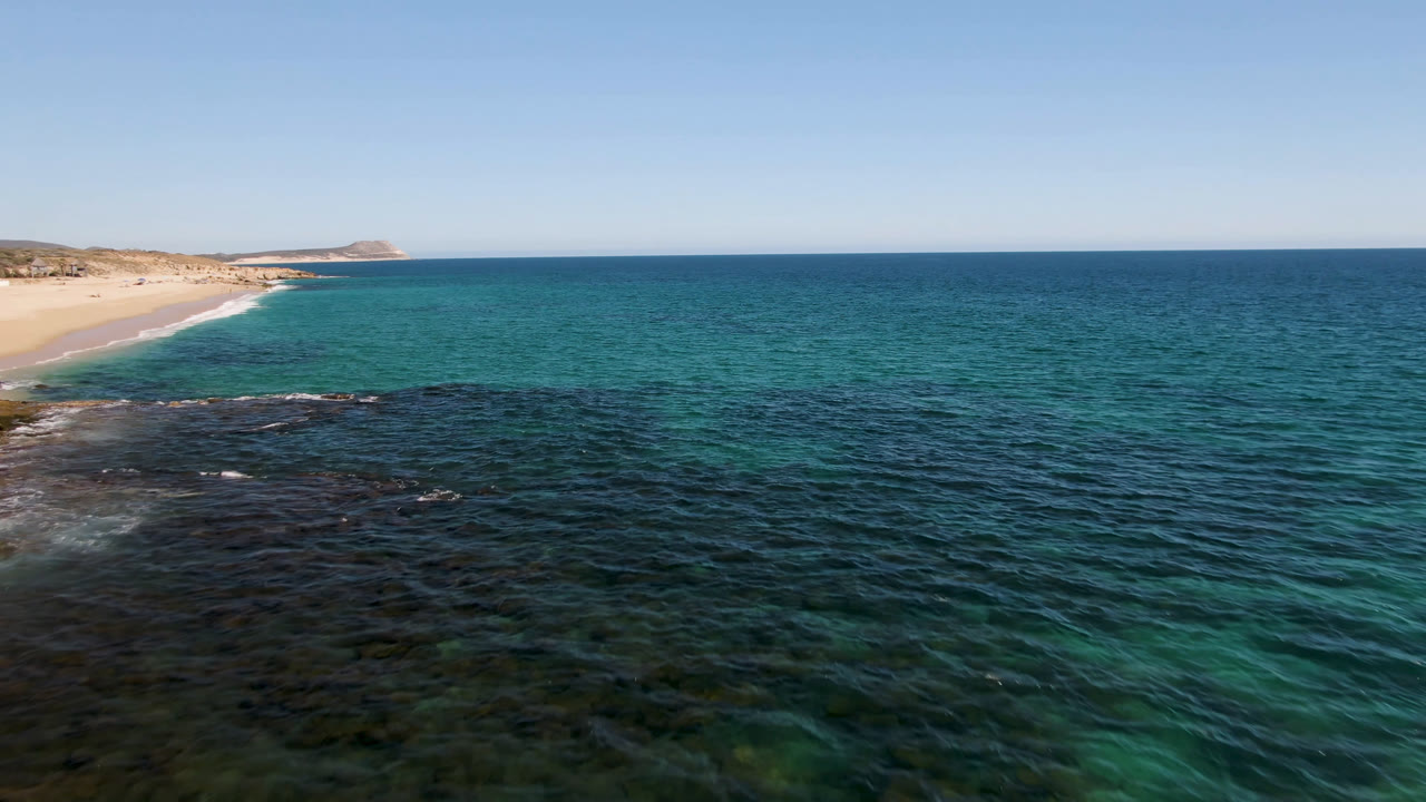 Aerial coastal view with turquoise water, dark reef patches, beach, and low hills.