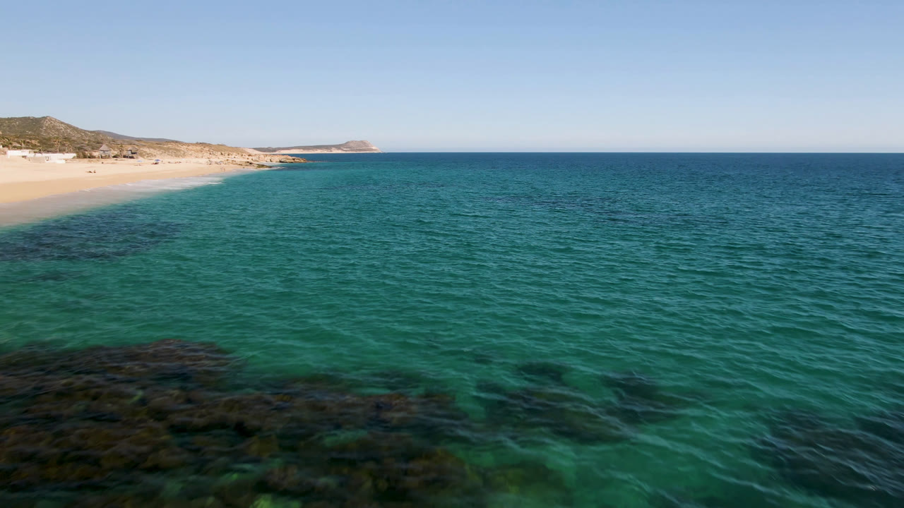 The second frame holds over clear green-blue water at Laguna Beach Los Cabos, with the sandy shore and low hills receding along the left side and no visible people.