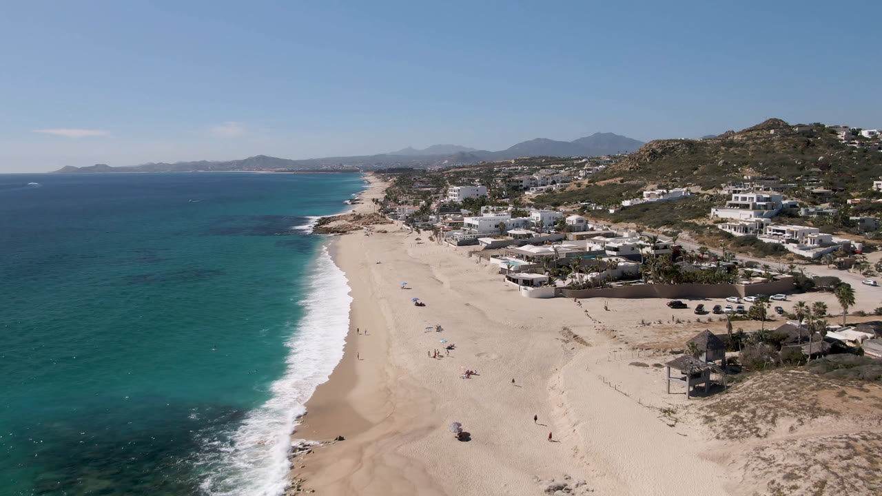 Aerial view of turquoise ocean, sandy beach, coastal houses, and hills.