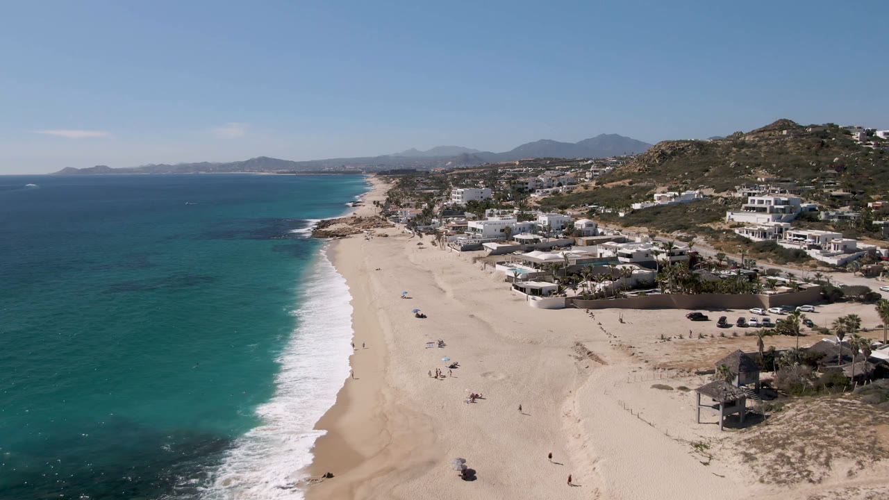 Laguna Beach in Los Cabos appears from above with a long surf line, open sand, white and tan buildings near the shore, and no identifiable people visible.