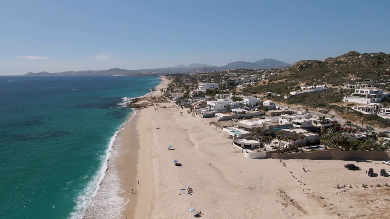 Opening aerial frame over Laguna Beach Los Cabos, Mexico, with turquoise water on the left, pale sand below, white beachside homes to the right, and no clearly visible people.