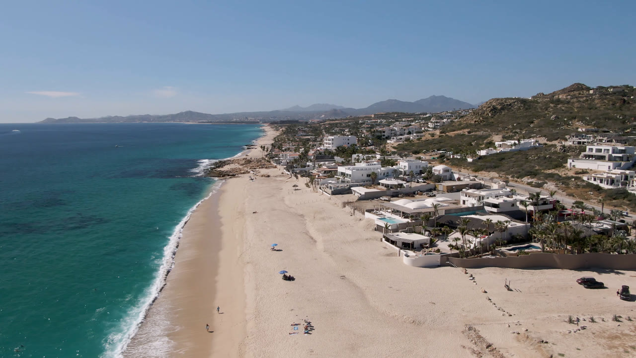 Aerial frame at 0:01 over Laguna Beach Los Cabos, Mexico, showing the surf line running diagonally beside beachside homes and open sand with no clearly visible people.