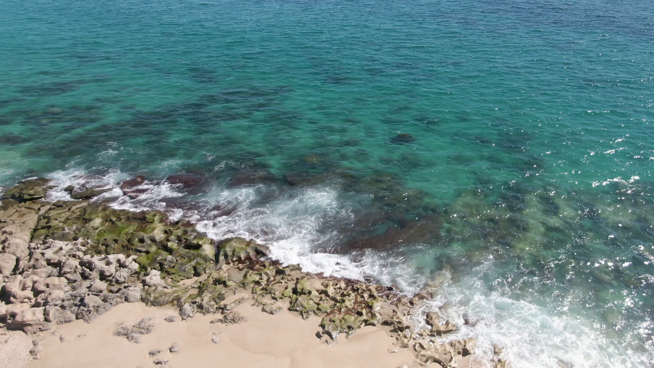Aerial frame over Laguna Beach Los Cabos showing turquoise water meeting dark rocks and pale sand, with white surf at the shoreline and no visible people.