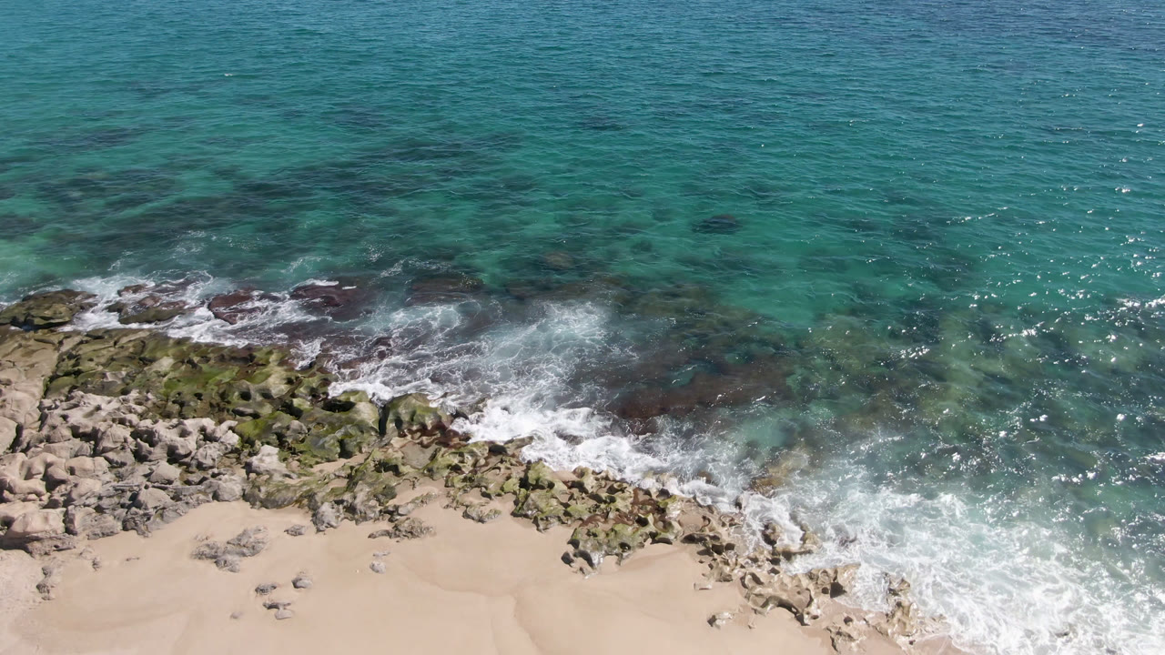Overhead Laguna Beach Los Cabos view with a broader burst of white water crossing the rocks and washing toward the sand, with no visible people.