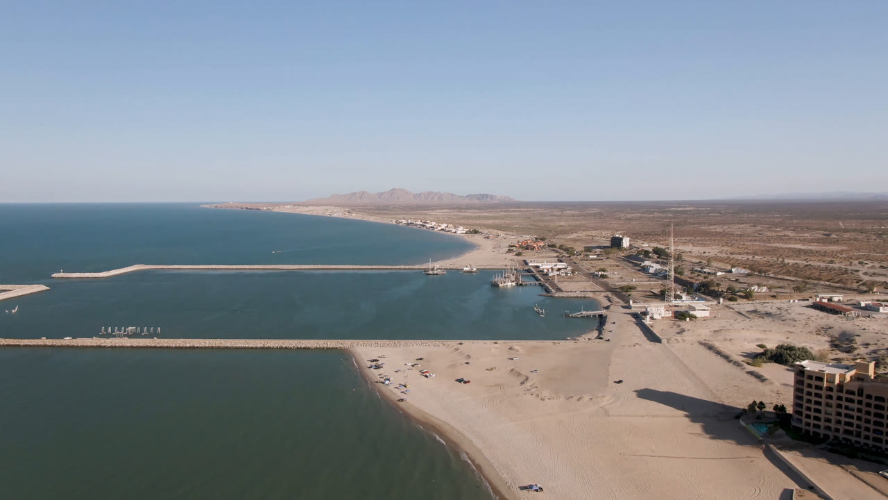 Aerial frame over San Felipe, B.C., Mexico with the pier crossing the water, the beach in the foreground, waterfront buildings at right, desert inland, and no visible people.