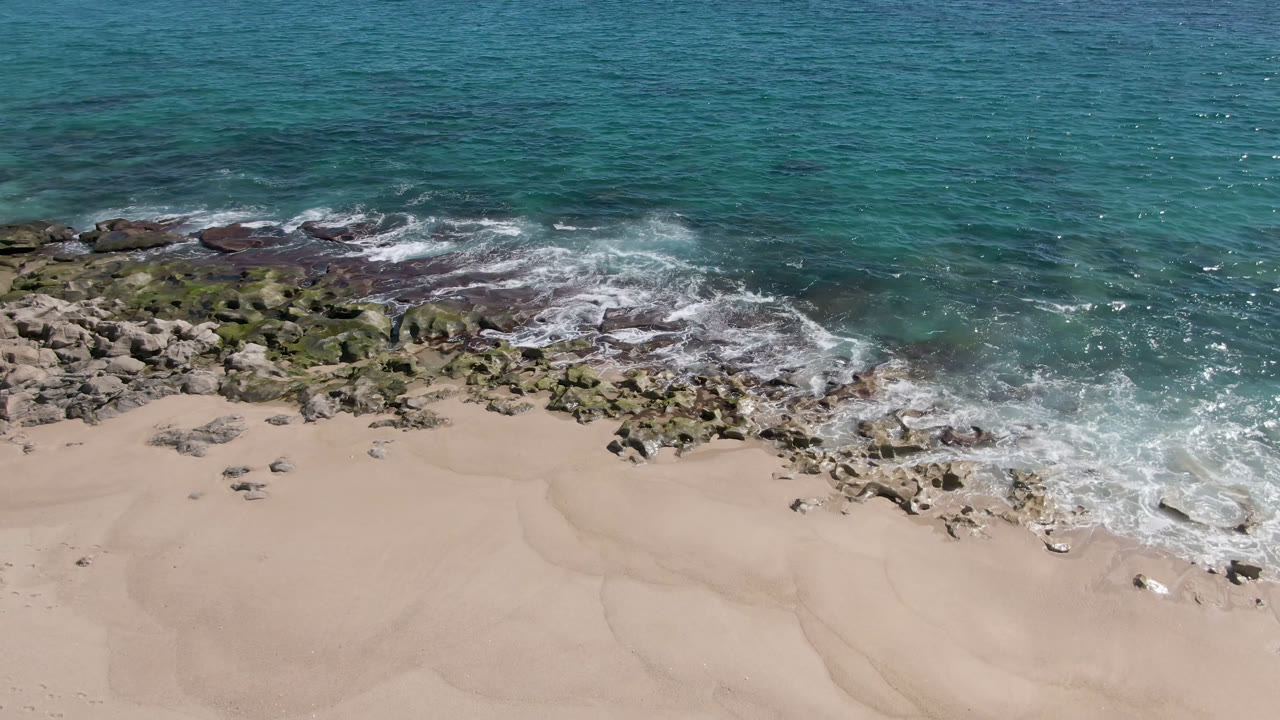 Overhead view of Laguna Beach Los Cabos with turquoise water breaking over dark rocks, tan sand below, and no visible people.