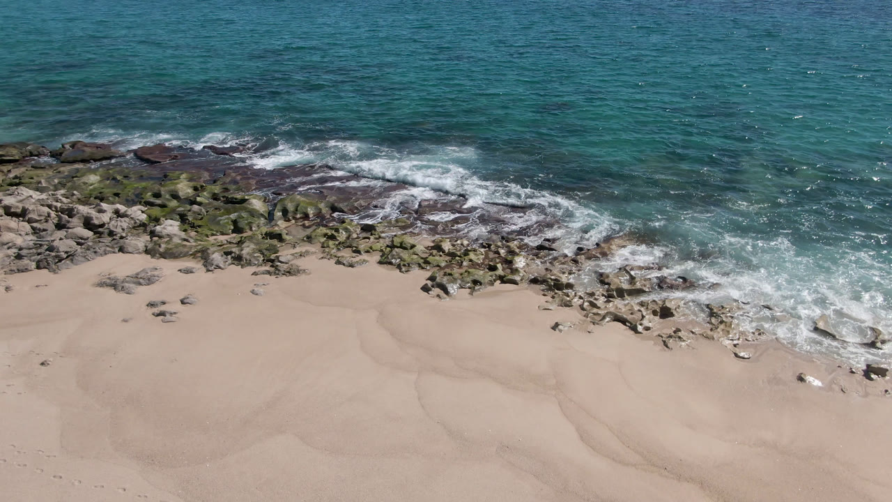 Aerial frame of Laguna Beach Los Cabos where white foam spreads along the rocky edge above a broad, empty stretch of sand with no visible people.