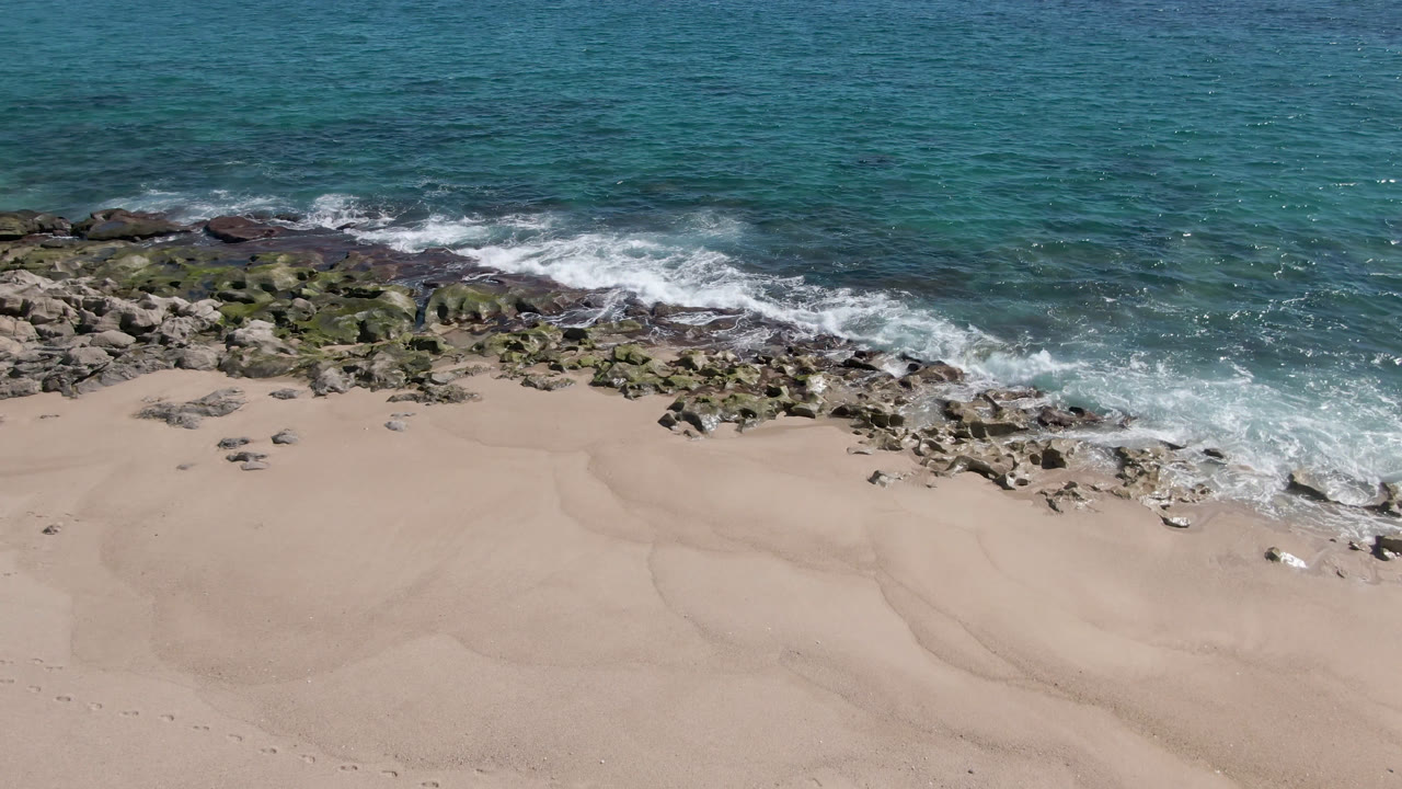 Drone view of rocky surf line and pale beach under bright daylight.