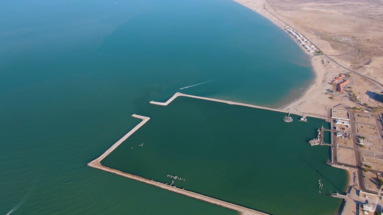 Aerial frame over San Felipe, Baja California, Mexico showing angular breakwaters enclosing green-blue water, a dock area, a small boat, sandy shoreline, and no visible people.