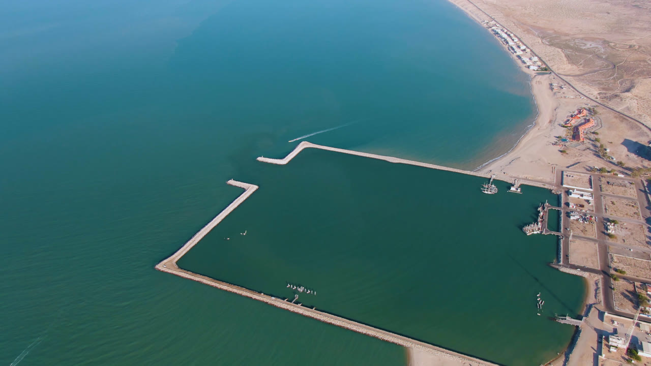 Opening aerial frame over San Felipe, Mexico, showing teal water around angular pale breakwaters, a sandy beach at the bottom edge, shoreline buildings to the right, and no visible people.