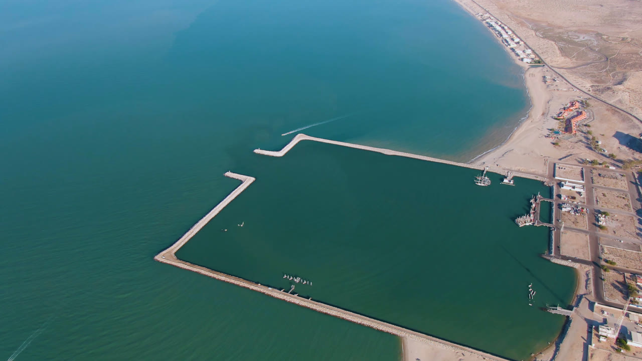 Second aerial frame over San Felipe, Mexico, with the breakwater basin centered, a small boat near the shore side, dry land along the upper right, and no visible people.