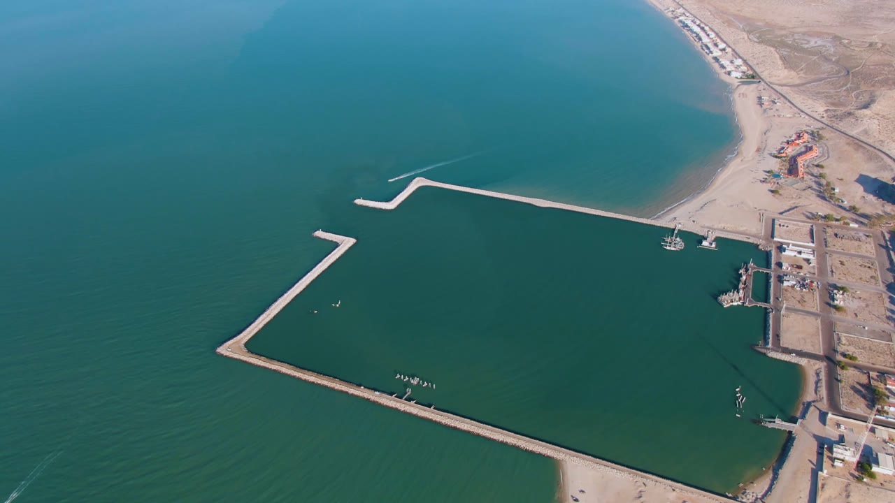 Drone view of a coastal basin with long breakwaters, beach, boats, and dry shoreline land.