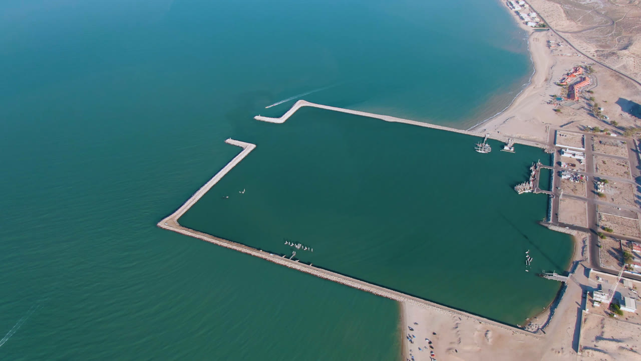 Opening aerial frame over San Felipe, Baja California, Mexico, with green open water, pale harbor breakwaters, sandy beach at the bottom, a small boat near the shore, and no visible people.