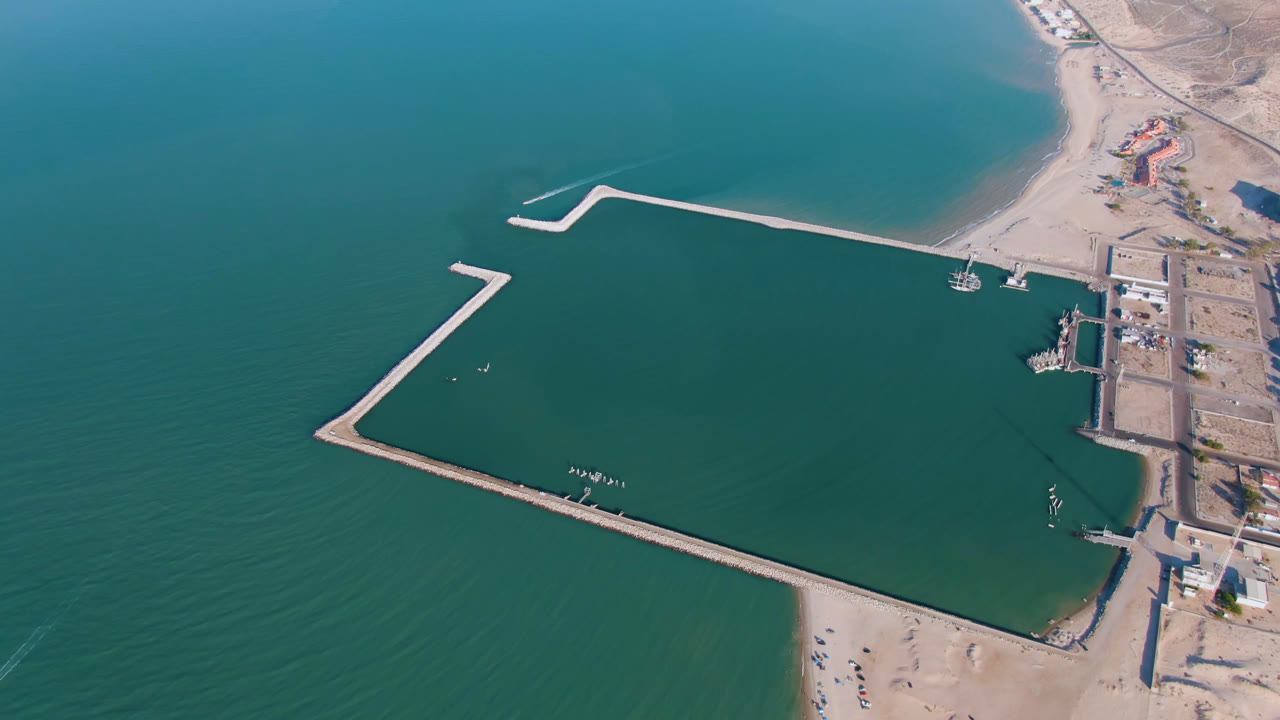 Aerial view over San Felipe, Baja California, Mexico, with the harbor basin centered, a long breakwater crossing the upper frame, beach and dry coastal lots on the right, and no visible people.