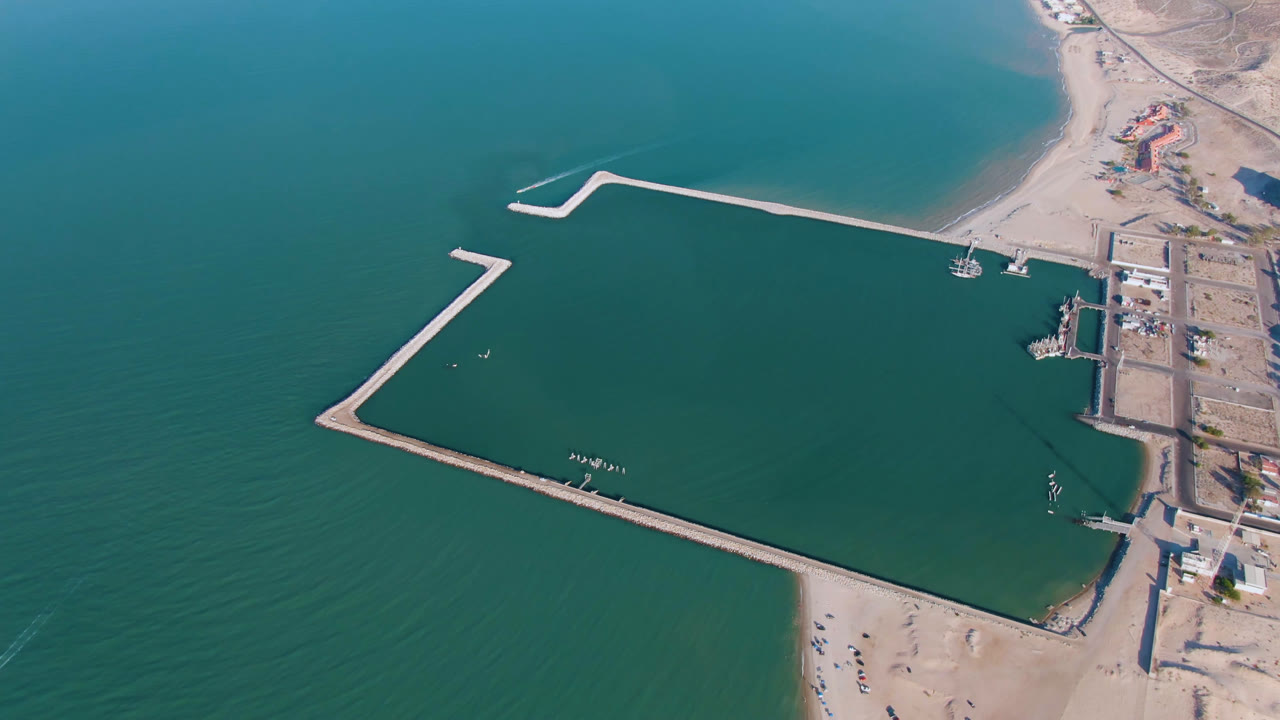 Drone view of a coastal harbor with angular breakwaters and a small boat by the beach.