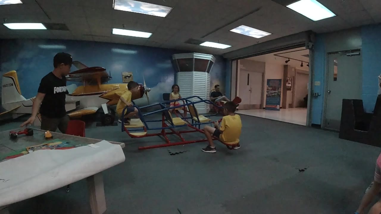 A wide view holds on children around the slanted indoor climbing structure, with the black-shirted child at left and a seated child near the right rail.