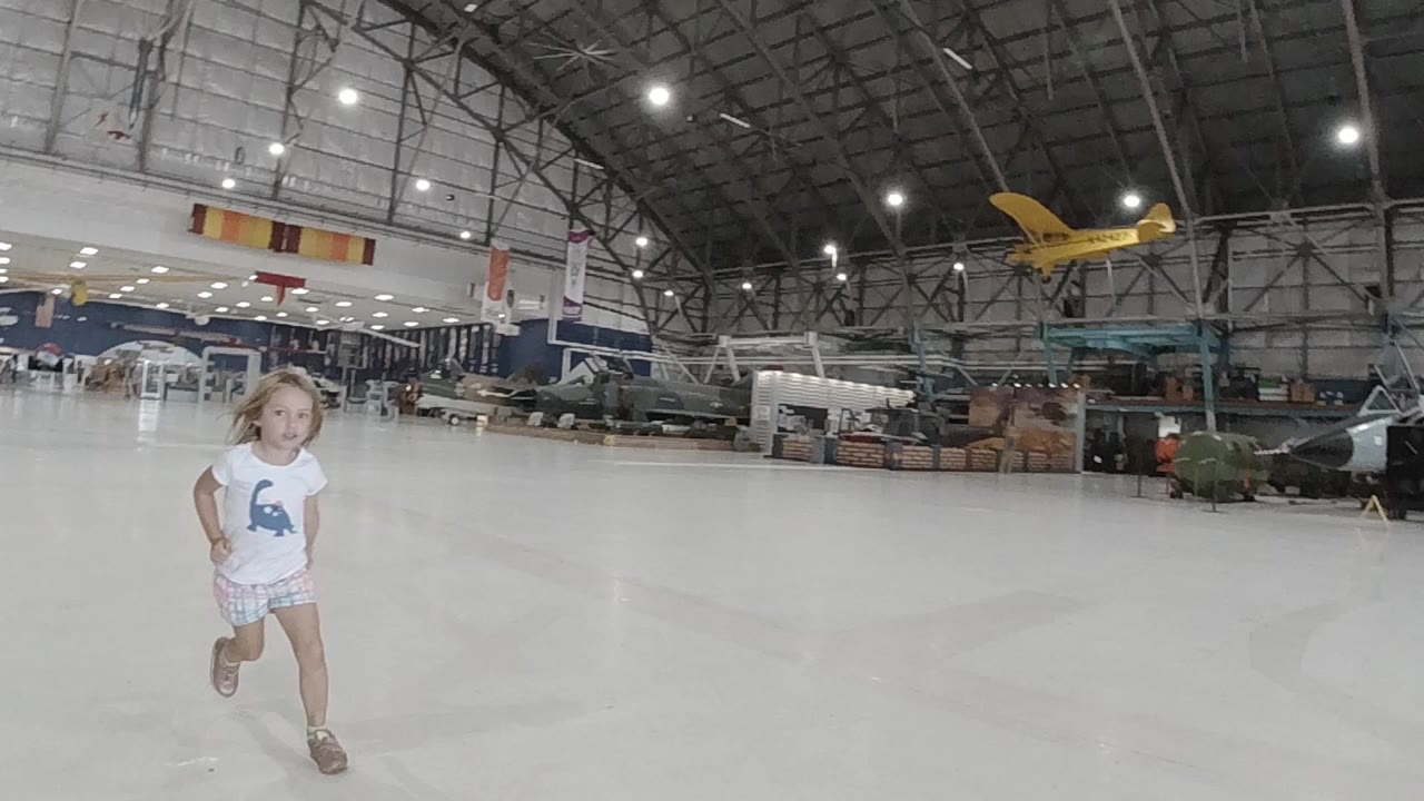 A young girl runs from the left side of a wide Colorado airplane hangar, with parked aircraft and a yellow plane visible under bright ceiling lights.