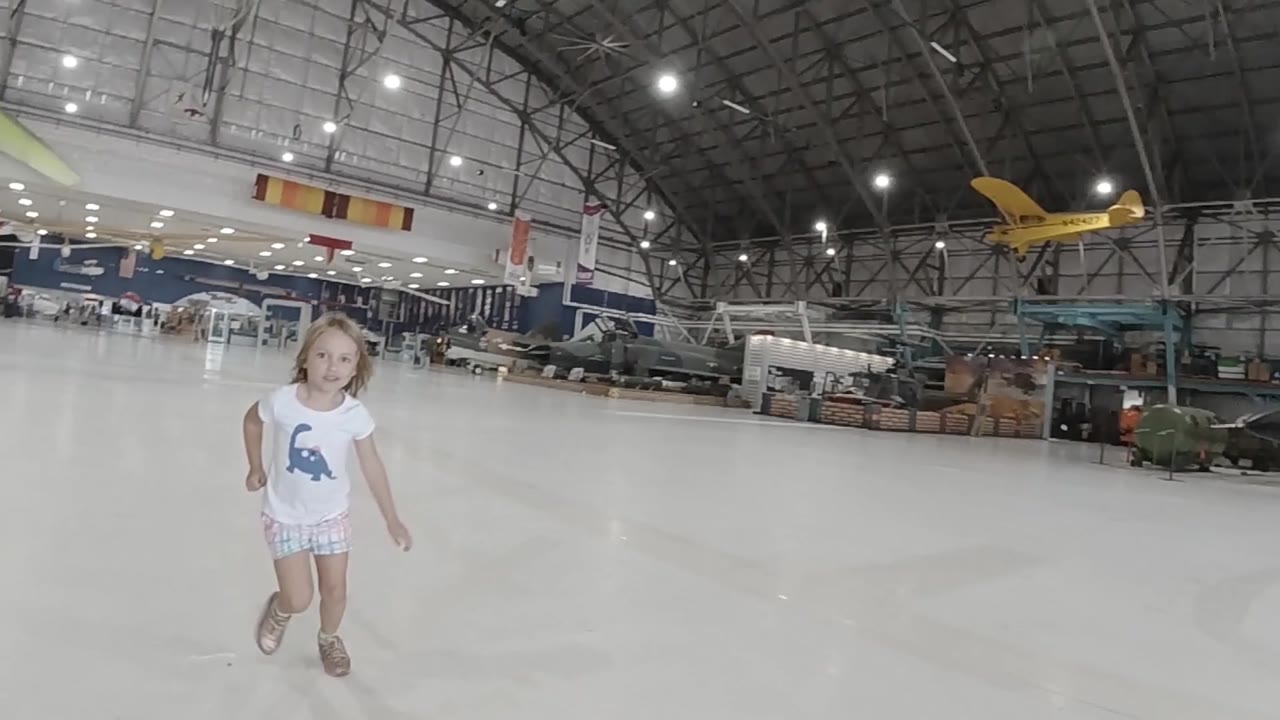 A smiling girl runs toward the camera in the open Colorado hangar, with the yellow plane suspended or parked high in the background and aircraft along the wall.