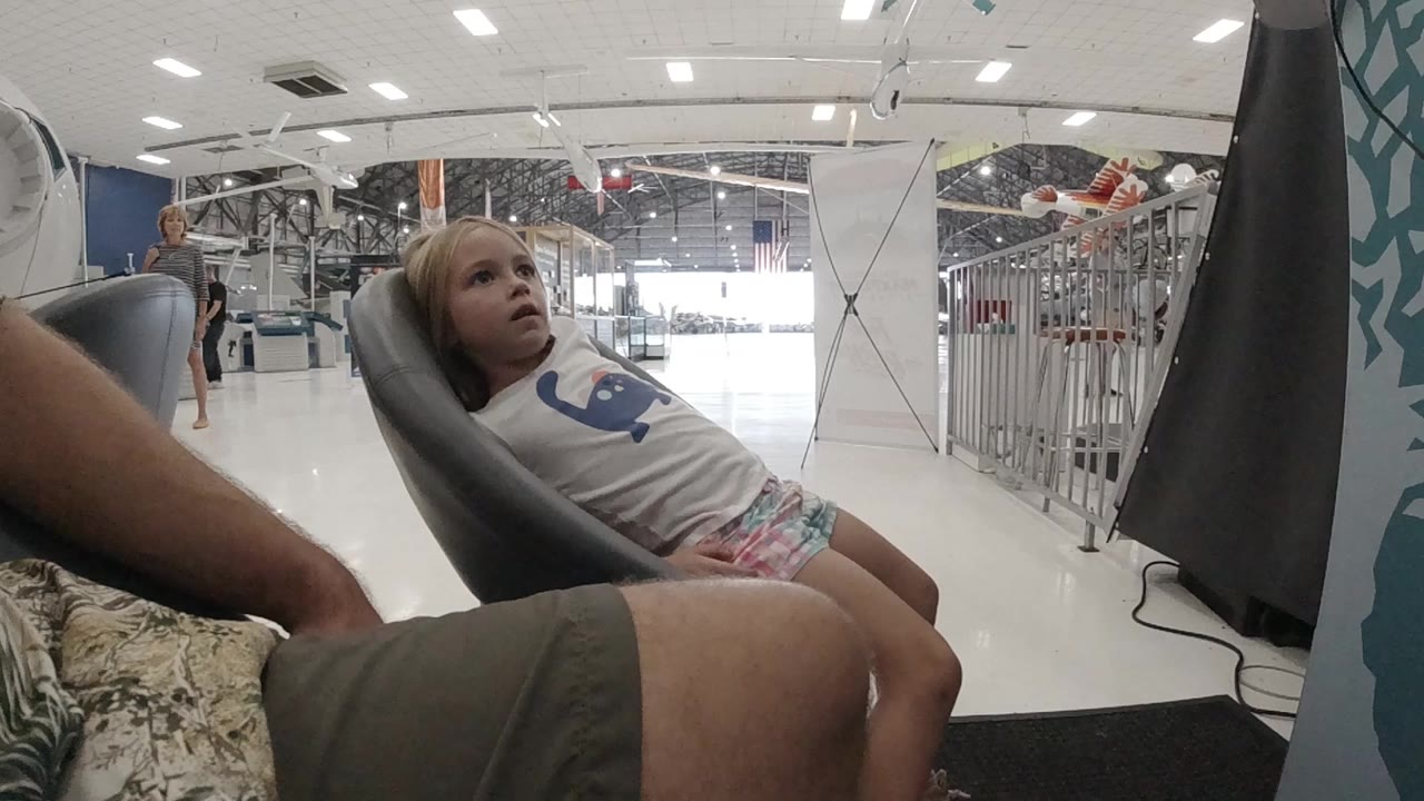 A blonde toddler girl reclines sideways in a dark gray egg-shaped lounge chair in a bright Colorado interior, with an adult leg in the foreground and metal barriers behind her.