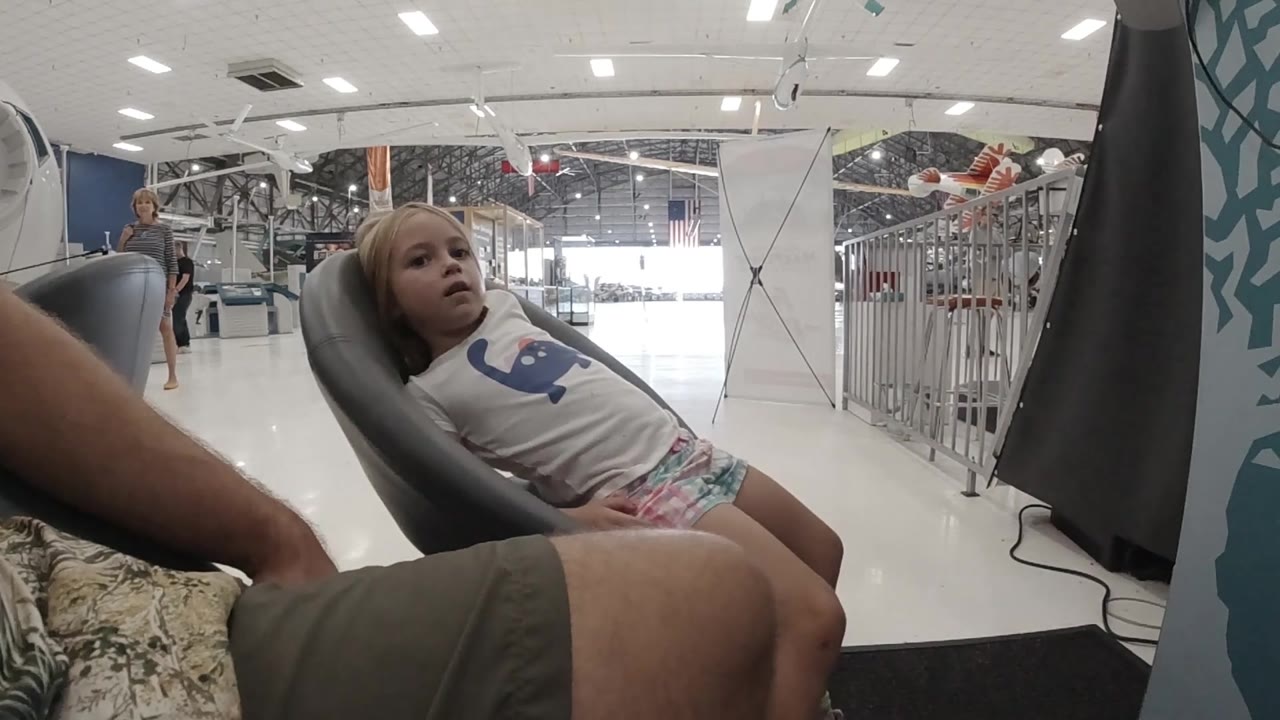 A low wide view shows the blonde toddler reclined in the chair, with a bright entry area, white flooring, and metal barriers behind her in Colorado.