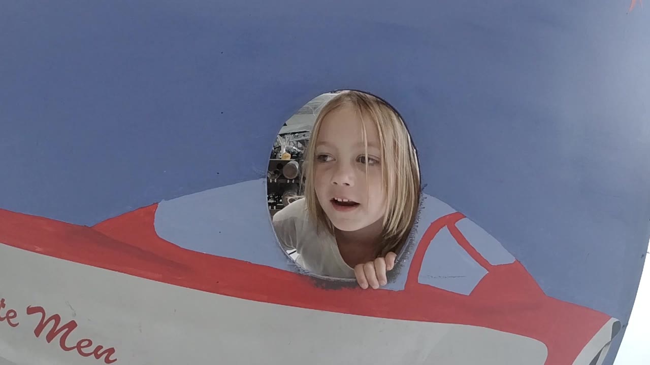 A blonde child in Colorado looks through a circular opening in a blue and red cardboard airplane display, with her fingers on the lower edge.