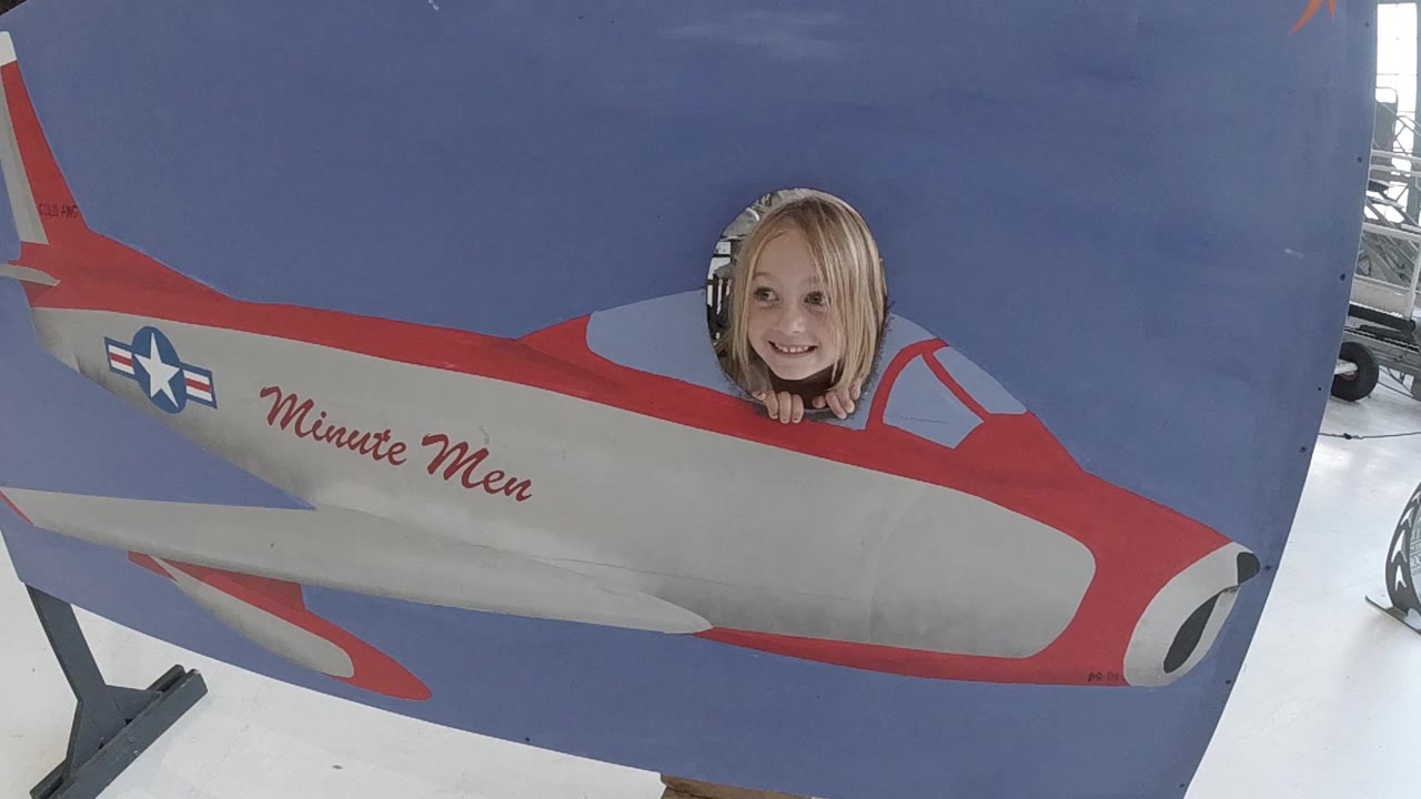At the start of the Colorado clip, a blonde woman smiles through the round cockpit hole of a red, white, and blue Minute Men airplane cutout inside a bright indoor space.
