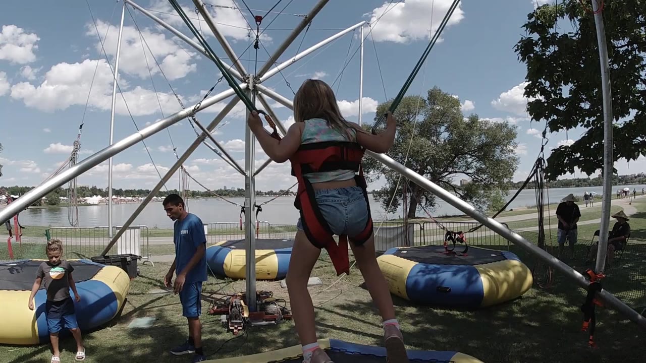 A harnessed jumper is seen from behind at a Colorado outdoor bungee attraction, standing low in the frame beneath metal tower tubes while staff and spectators wait near blue and yellow inflatable pads.