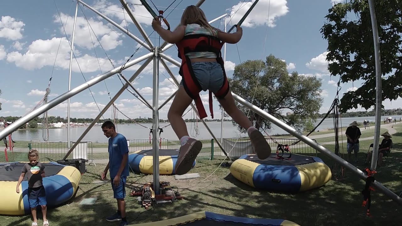 The jumper is higher in the bungee structure with legs extended outward, while staff and a child remain below near the blue and yellow inflatable landing pads.